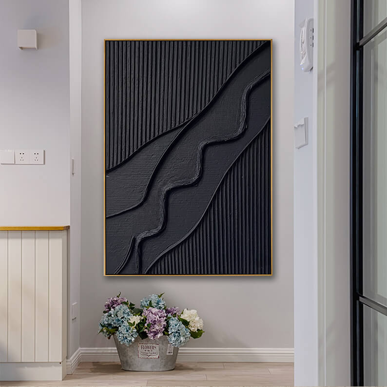 A home hallway or entryway featuring the black textured canvas art mounted on a smooth, light gray wall. On the light wood floor directly beneath the painting is a rustic galvanized metal bucket filled with vibrant blue, white, and purple hydrangea flowers. To the left is a white beadboard cabinet with a wood countertop, and to the right is an open door frame, creating a bright and welcoming transition space.