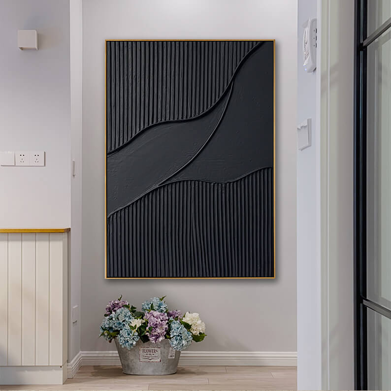 A bright, homey entryway scene. The matte black textured art piece is hung on a pale gray wall. On the light wood floor directly beneath the painting is a rustic metal garden bucket labeled "FLOWERS" filled with a lush bouquet of blue, purple, and white hydrangeas. A white door frame is visible on the right, and a cottage-style white wood panel cabinet is on the left.