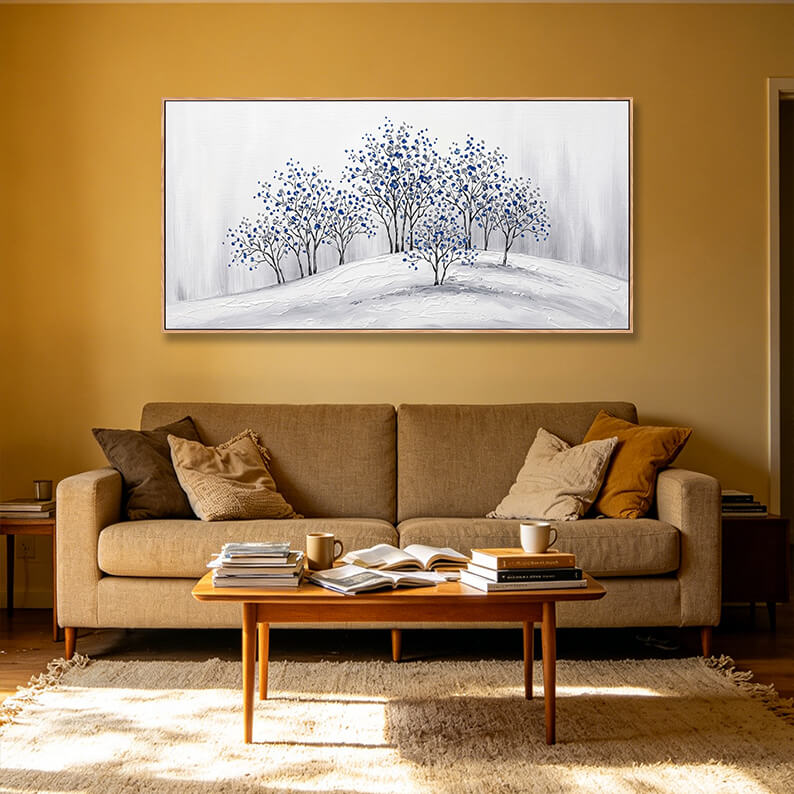 Panoramic horizontal blue and white tree grove painting hanging on a warm yellow wall above a tan fabric couch.