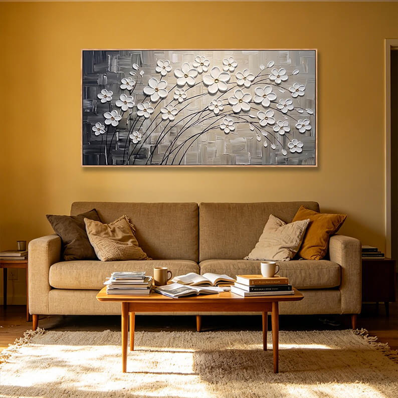 Panoramic horizontal white blossom painting styled over a tan sofa in a living room with warm golden-yellow walls.