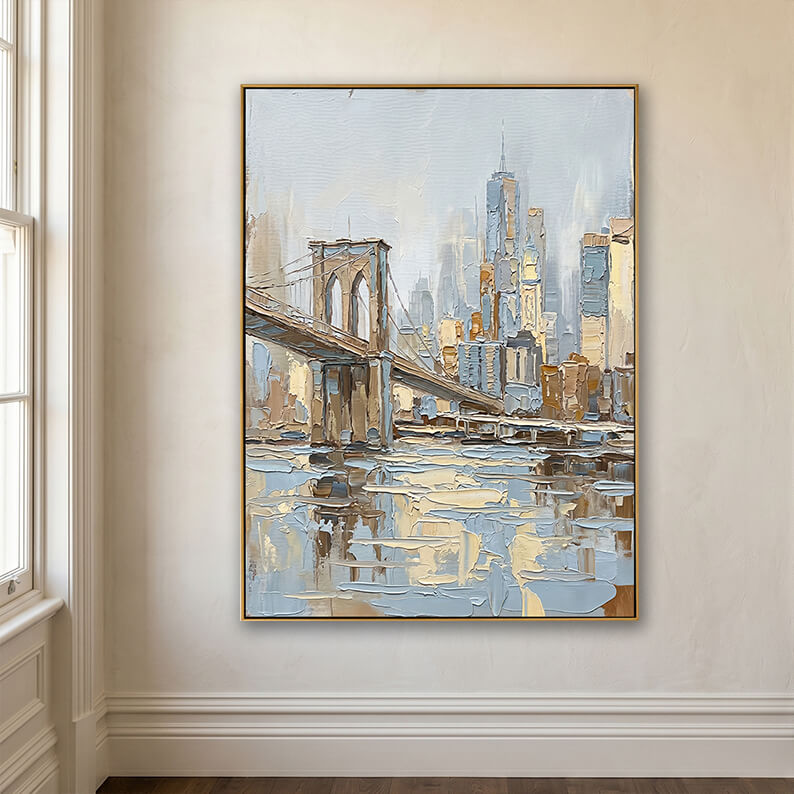 Framed textured city skyline painting depicting the Brooklyn Bridge, mounted on a minimalist cream-colored plaster wall. The corner of a white-paneled window frame is visible on the left. The bottom of the wall features elegant white baseboard trim meeting dark wood floors.