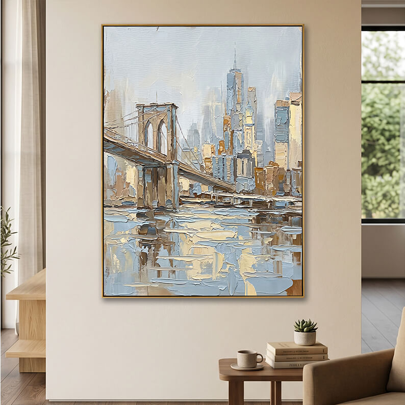A large, vertically oriented impasto canvas painting of the Brooklyn Bridge and Manhattan skyline in a gold frame, hung on a warm beige wall. Below the painting, a low wooden side table holds a stack of books, a small potted succulent, and a coffee mug. To the left, a window is dressed with sheer white curtains, casting soft natural light across the scene.