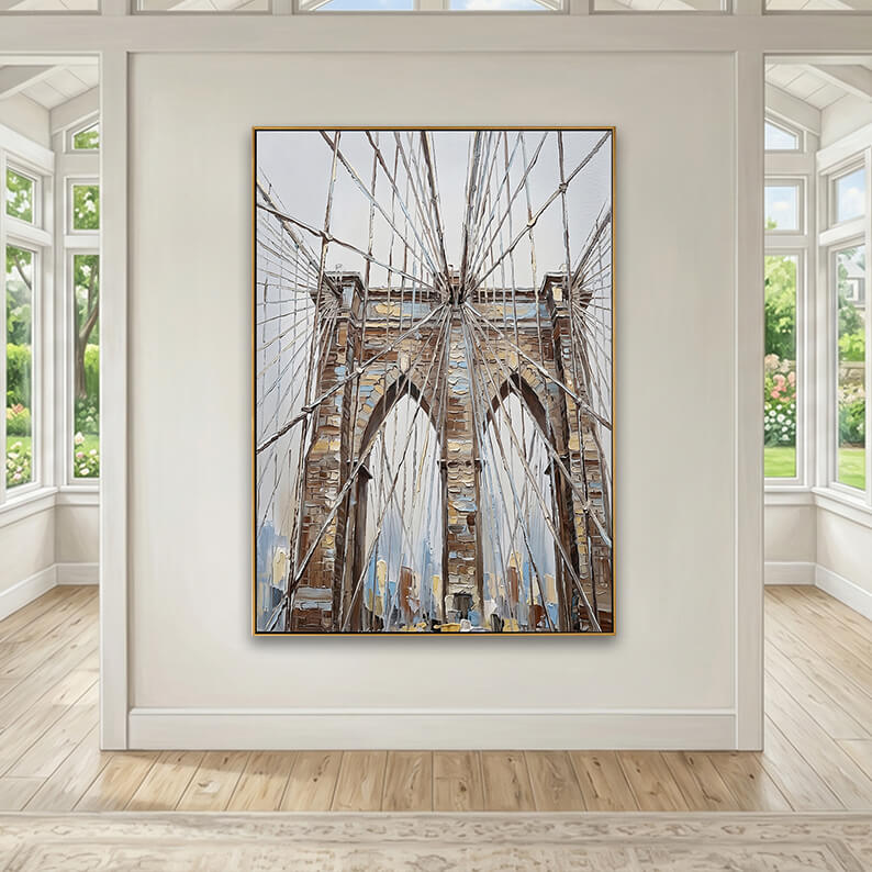 A spacious interior room mock-up showcasing the gold-framed Brooklyn Bridge painting hung in the center of a symmetrical, off-white accent wall. The room is a bright, sunroom-style space with high ceilings and large paned windows on both the left and right walls, showing views of a lush green backyard garden. The floor is made of light-toned natural wood planks, and a classic neutral patterned area rug is visible in the foreground.