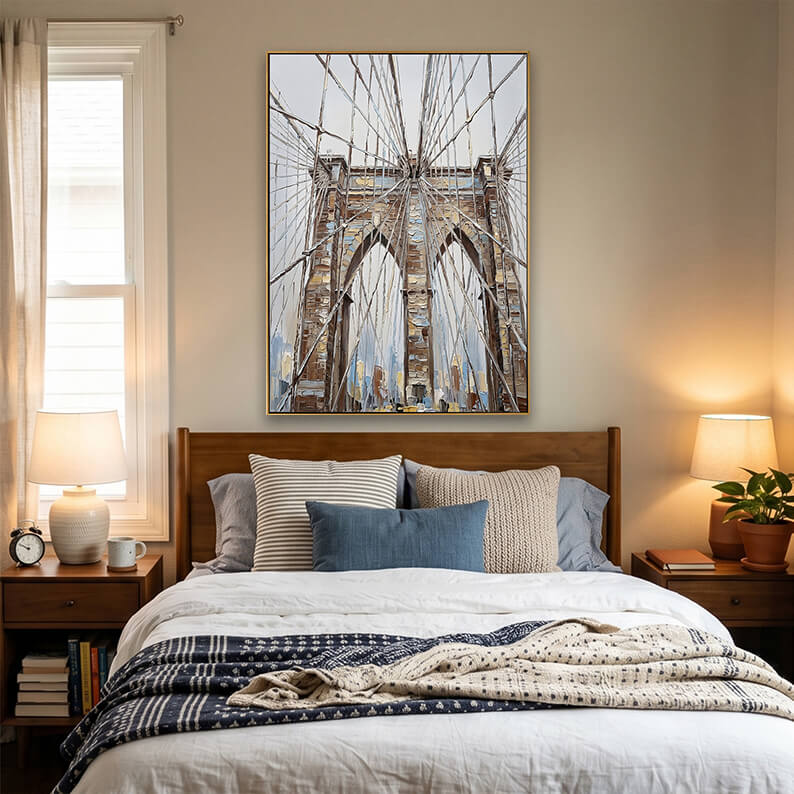 A warm bedroom interior setup. The gold-framed Brooklyn Bridge artwork is displayed on a beige wall above a solid, dark wood headboard. The bed is styled with clean white bedding, a light blue accent pillow, and a heavy knit blue-and-white patterned throw blanket draped over the foot of the bed. Dark wood nightstands on either side hold matching bedside lamps with round ceramic bases and glowing fabric shades. A window with light filtering through beige curtains is visible on the left.