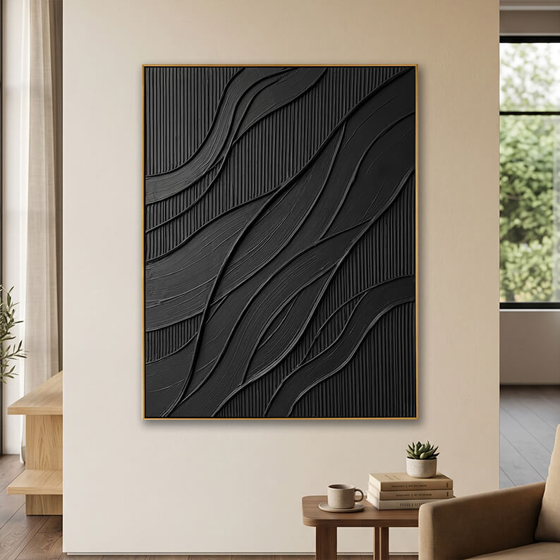 The textured black framed artwork hanging on a neutral-toned wall in a cozy modern room. In the foreground, a small wooden side table holds a ceramic mug and a stack of books topped with a tiny potted succulent. An armchair is partially visible on the right, and a bright window looking out to green trees is in the background.