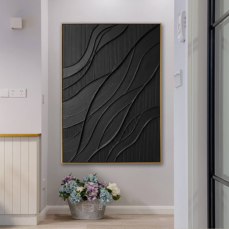 A vertical textured black art piece in a gold frame hanging in a gray-walled home entryway. Directly beneath the painting, a galvanized rustic metal bucket filled with blue, purple, and white hydrangeas rests on the light wood floor. White wood paneling and a sliding door frame are visible on the sides.