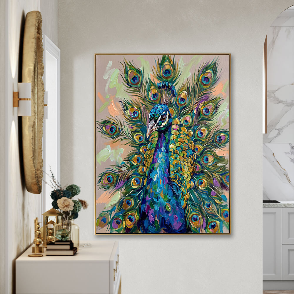 The vibrant peacock impasto painting is staged on a light gray, textured plaster wall in an elegant residential hallway. To the left, a white console table with gold-accented modern handles holds a stack of books, a small gold metal lantern, and a clear glass vase with white and blue flowers. Above the console table on the adjacent wall hangs a large, round decorative rustic gold frame mirror. To the right, a glimpse of a modern kitchen with a gray cabinet and white marble wall is visible.