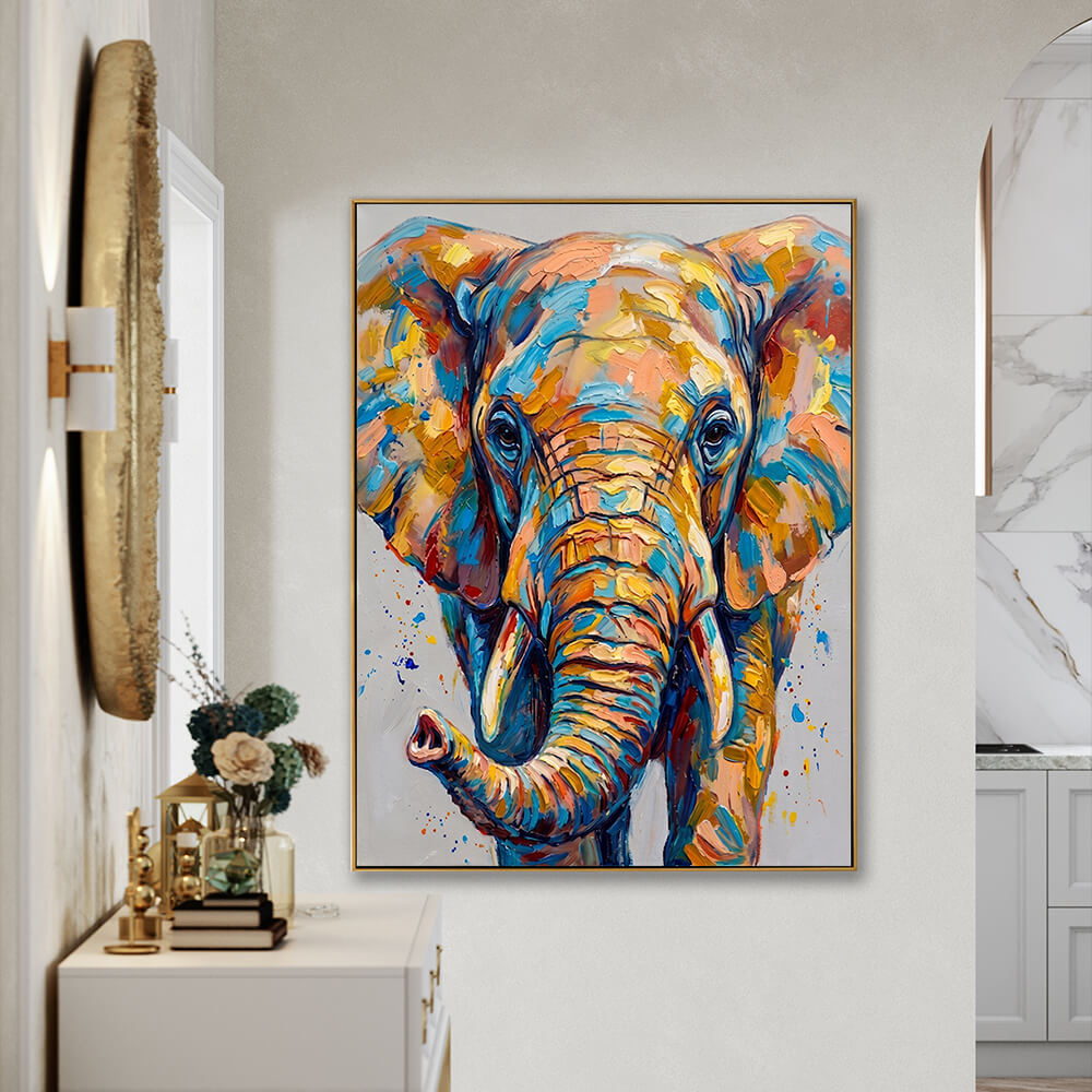 The colorful textured elephant painting, framed in gold, mounted on a warm beige wall in a modern entryway hallway. On the left, a large, textured round gold mirror with wall sconces hangs over a white console table decorated with books, a small lantern, and a floral vase. To the right, an arched walkway reveals a bright, clean kitchen with white cabinetry and marble tiling.