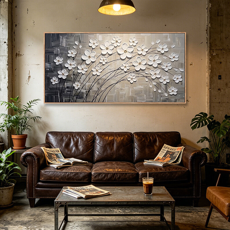 Panoramic white blossom wall art styled above a brown leather chesterfield sofa in a rustic loft living room with concrete walls.